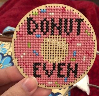 A cross stitch on a circular piece of wood of a pink sprinkle donut with the words Donut Even